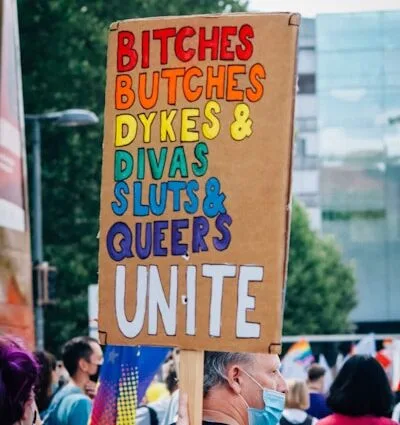 Protestplakat mit den Worten 'Bitches, Butches, Dykes, Sluts & Queers Unite' in bunten Buchstaben.